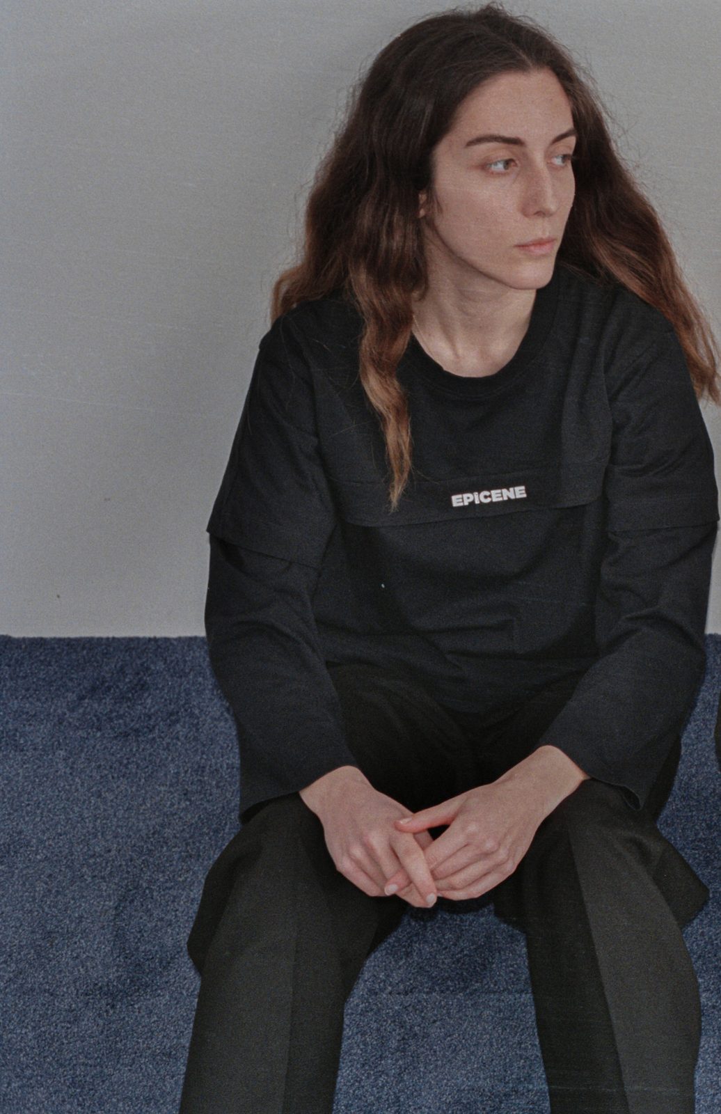 A model in a black Gender Fluid t-shirt and white trousers leaning forward in a stockroom