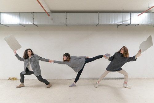 Three performers in a line holding sheets of paper, arms outstretched, in an empty concrete space