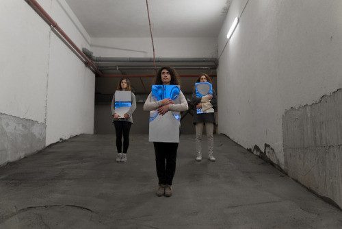 Three performers holding blue and white boards walking towards the camera in an underground car park