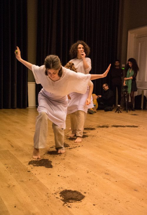 Two performers moving across a wooden floor scattered with soil