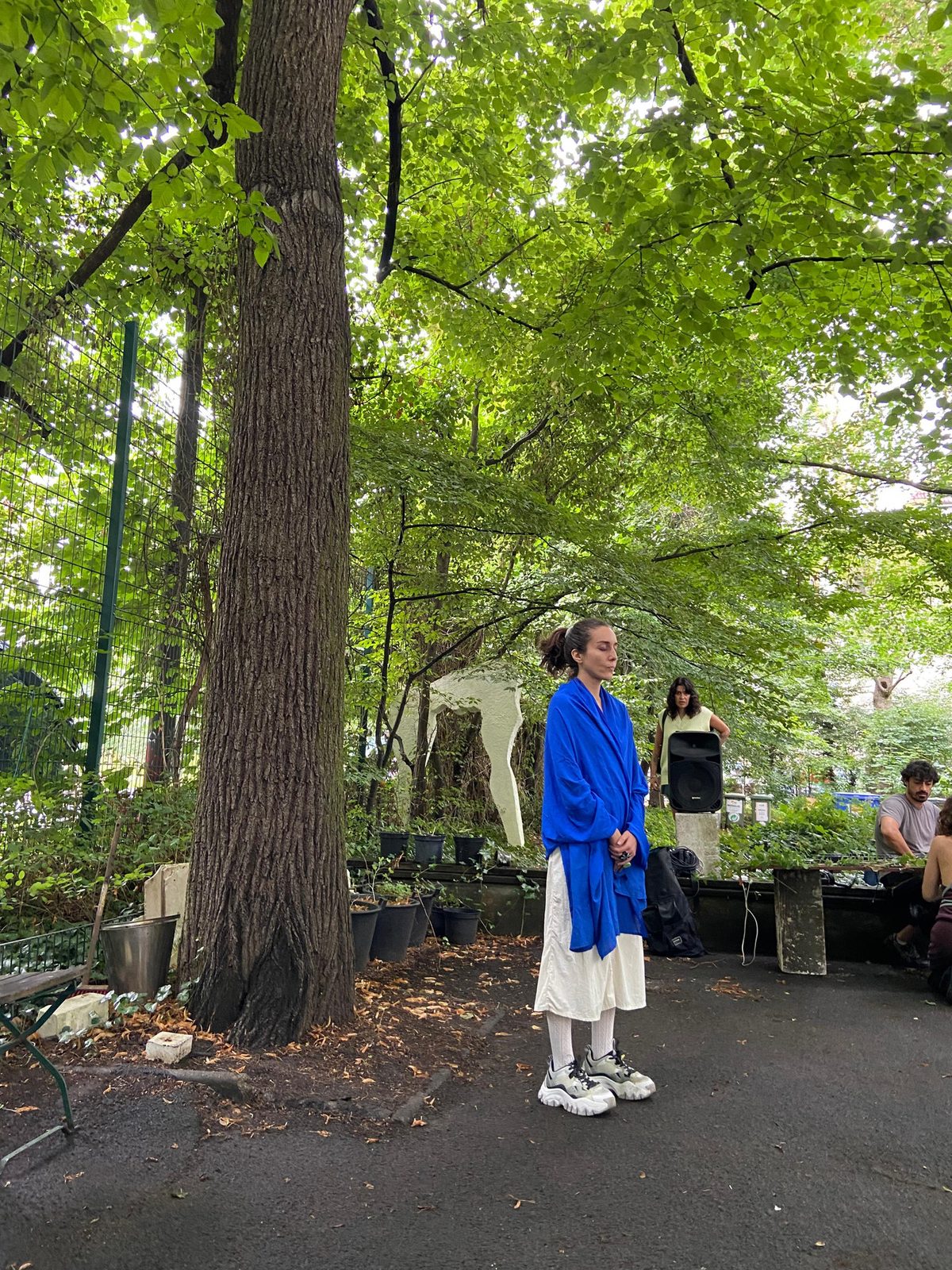 Dila in a blue jacket standing at the foot of a tall tree in the GlogauAIR courtyard, audience seated behind