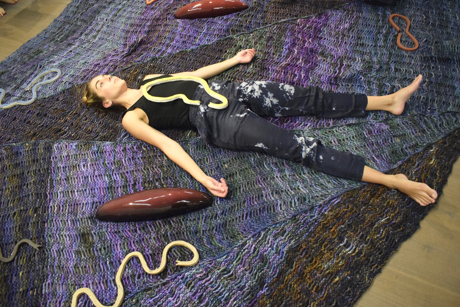A performer lying on a large knitted carpet in purple and blue tones, with rope and clay forms
