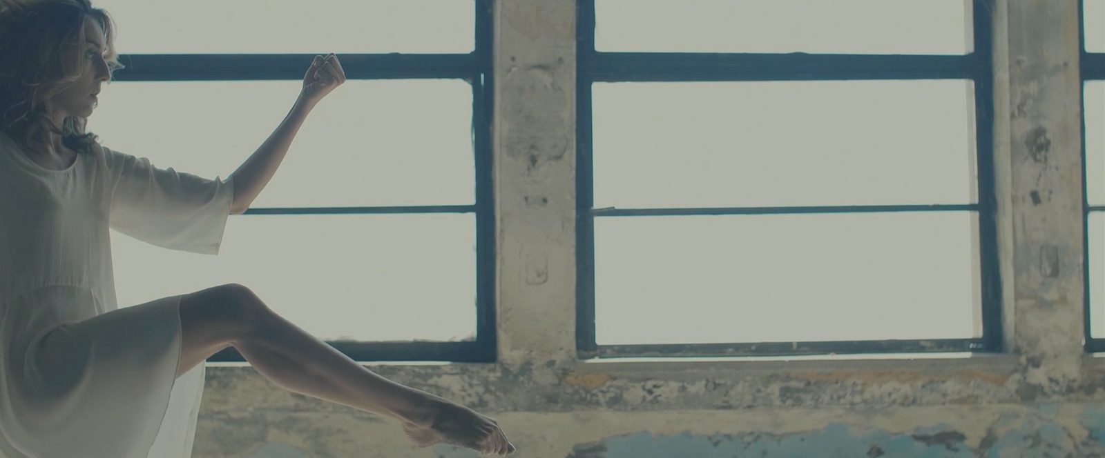 A dancer seated on a windowsill with one leg raised, still from the video