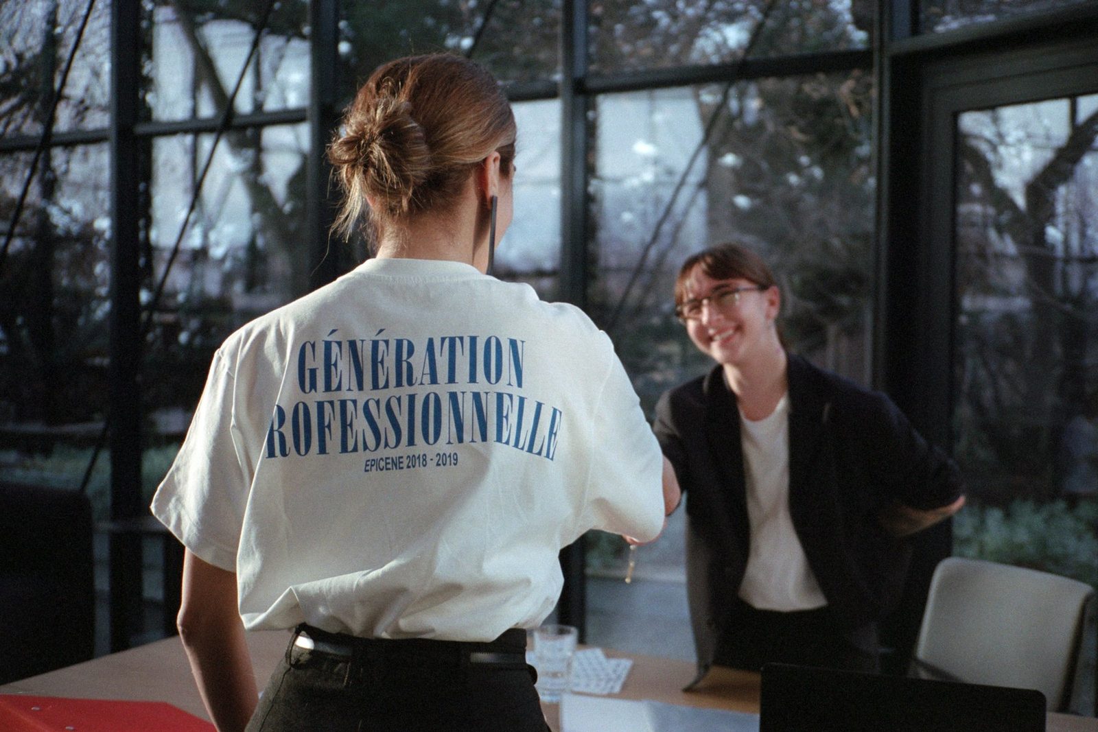 A model in a Génération Professionnelle t-shirt talking to a seated woman at a glass-walled office