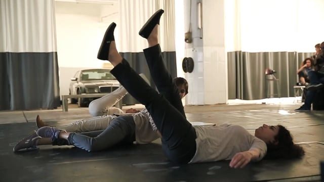 Two performers lying on their backs with legs raised, an audience seated along the wall