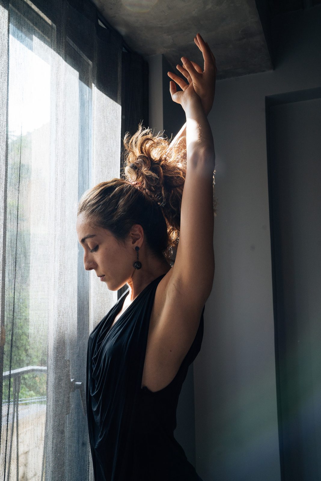 Dila with one arm raised beside a curtained window