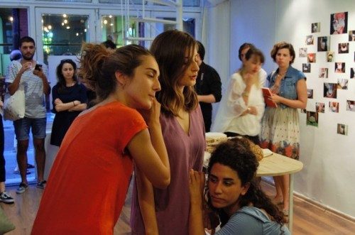 Three performers leaning together in a gallery, visitors looking on
