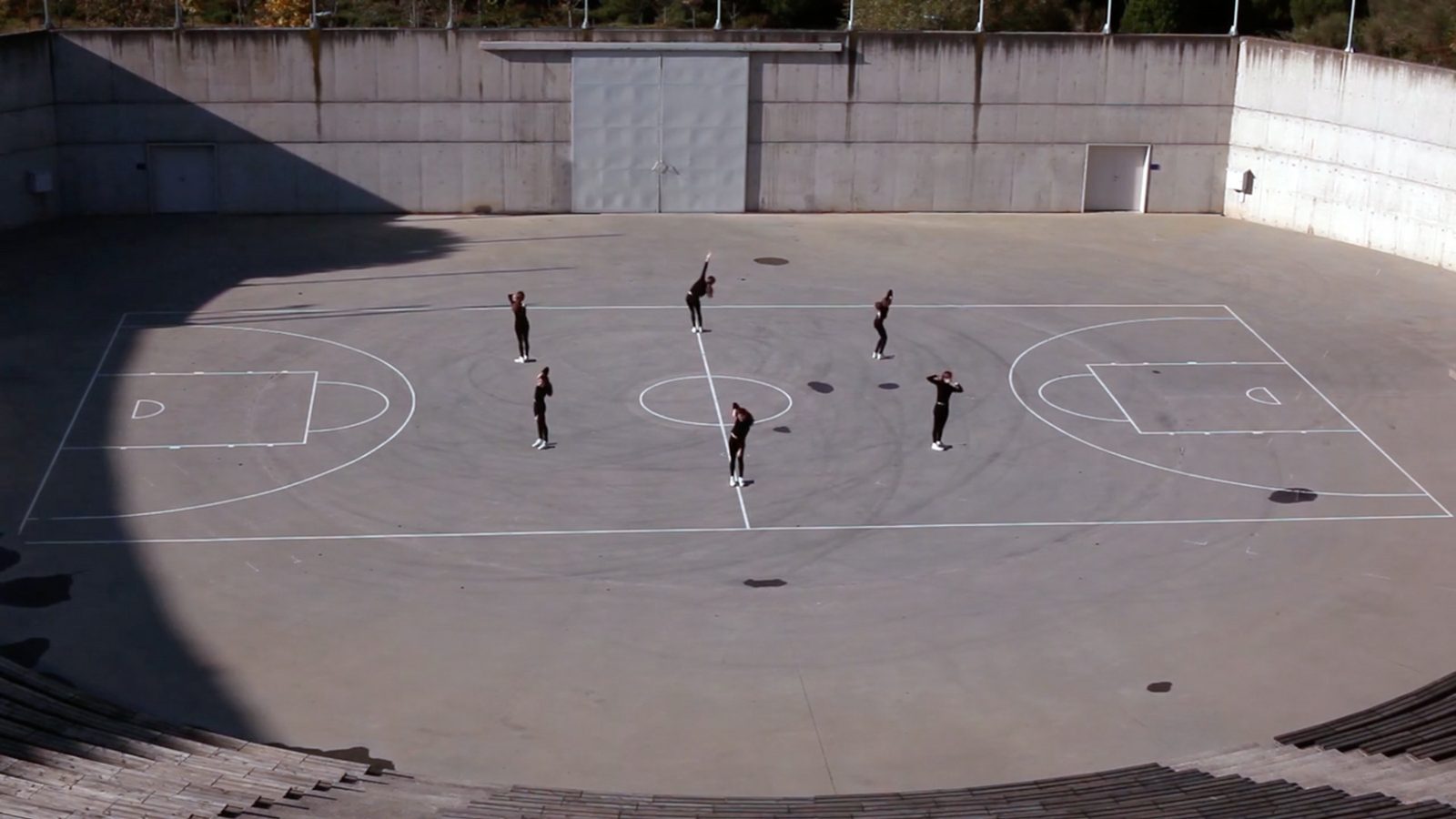Five dancers spread across an empty concrete basketball court seen from above