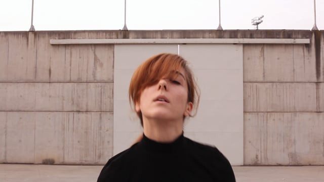 A dancer in black tilting her head back in front of a concrete wall, still from the video