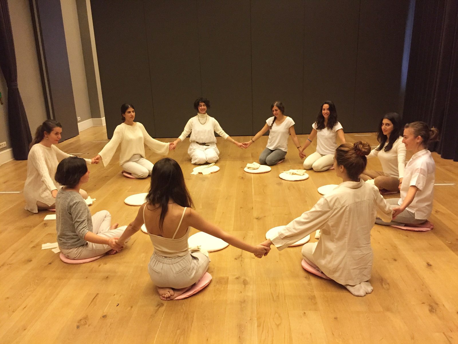 Participants dressed in white seated in a circle on a wooden floor, holding hands