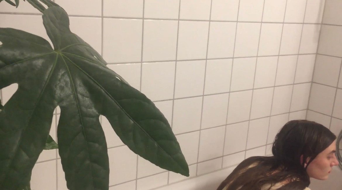 A woman lying in a bathtub beside a large leaf against white tiles