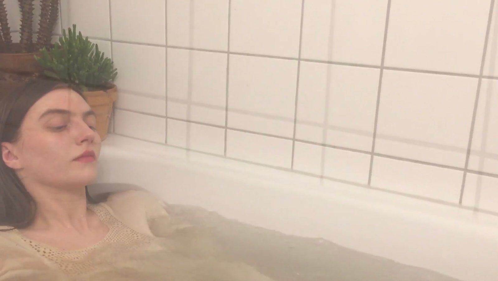 A woman lying in the bath with her eyes closed, a plant on the ledge behind her