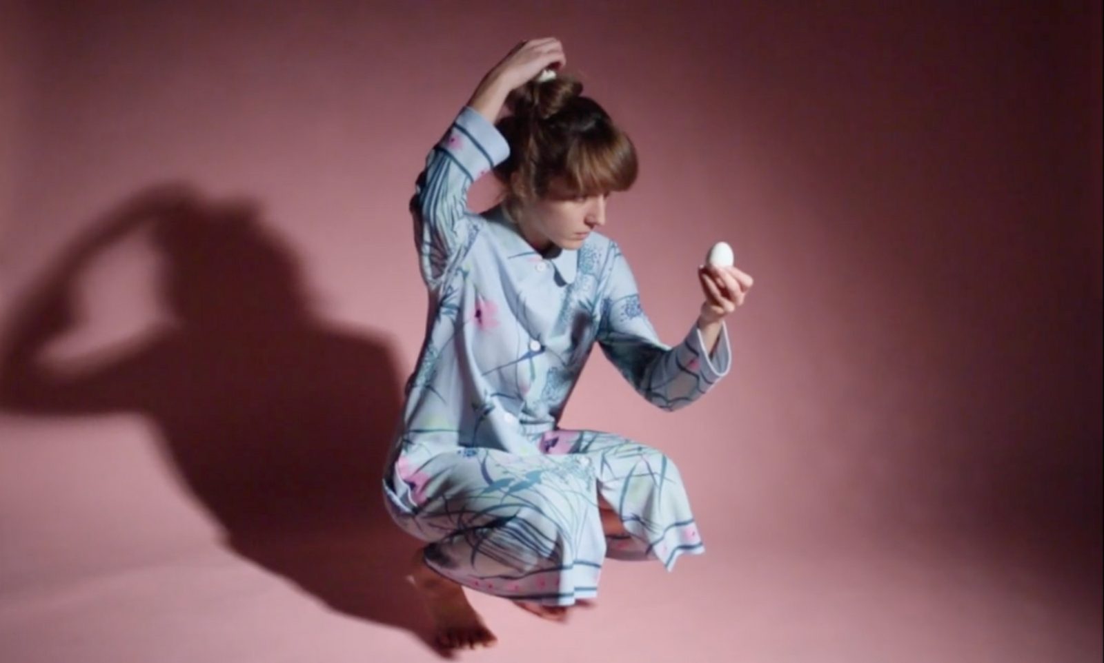 Dila in pyjamas crouched on a pink floor, holding an egg above her head